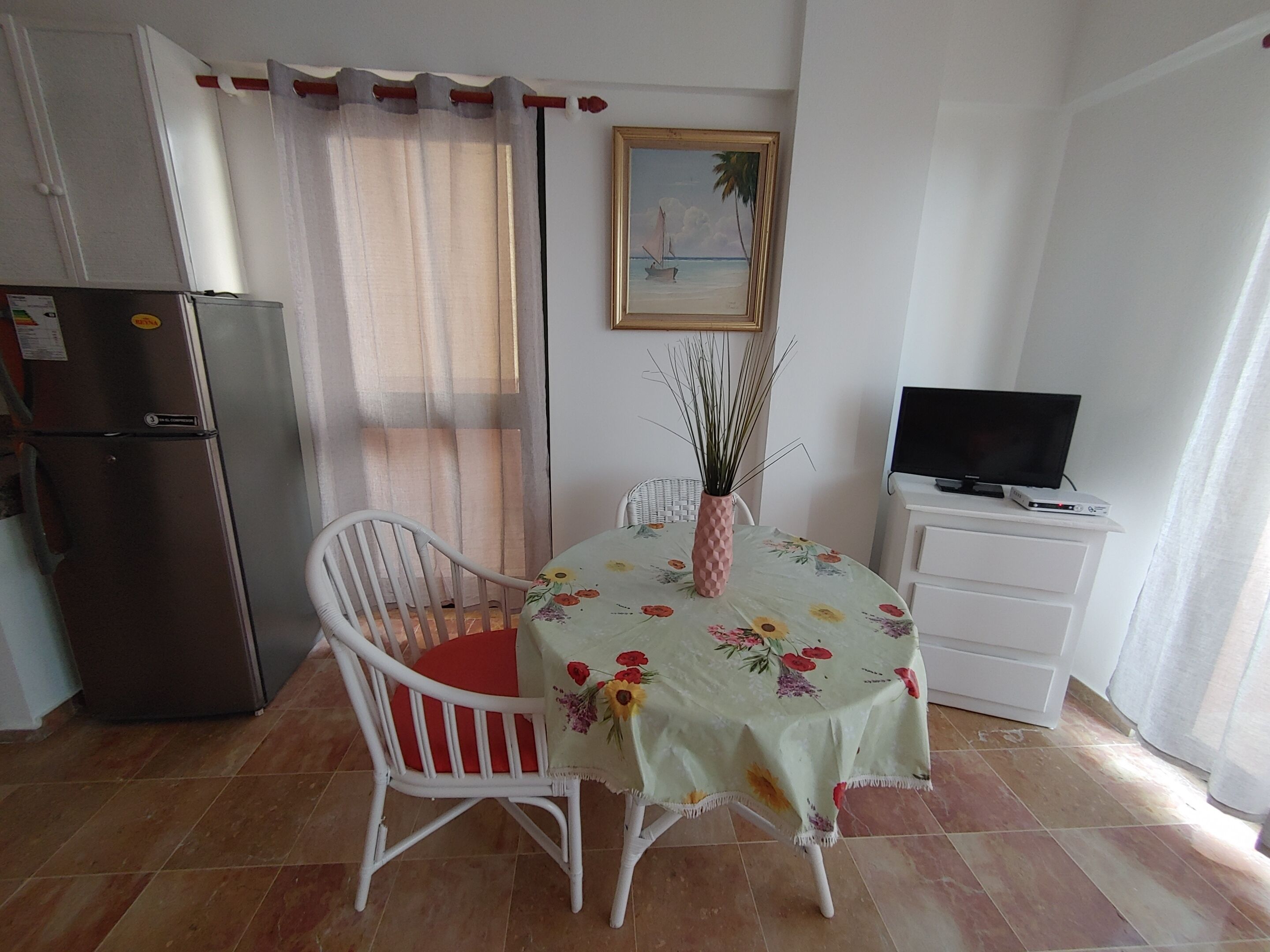 A small dining area with a table covered in a floral tablecloth, a white chair with a red cushion, a vase with plants, a refrigerator, a television on a stand, and a painting on the wall.