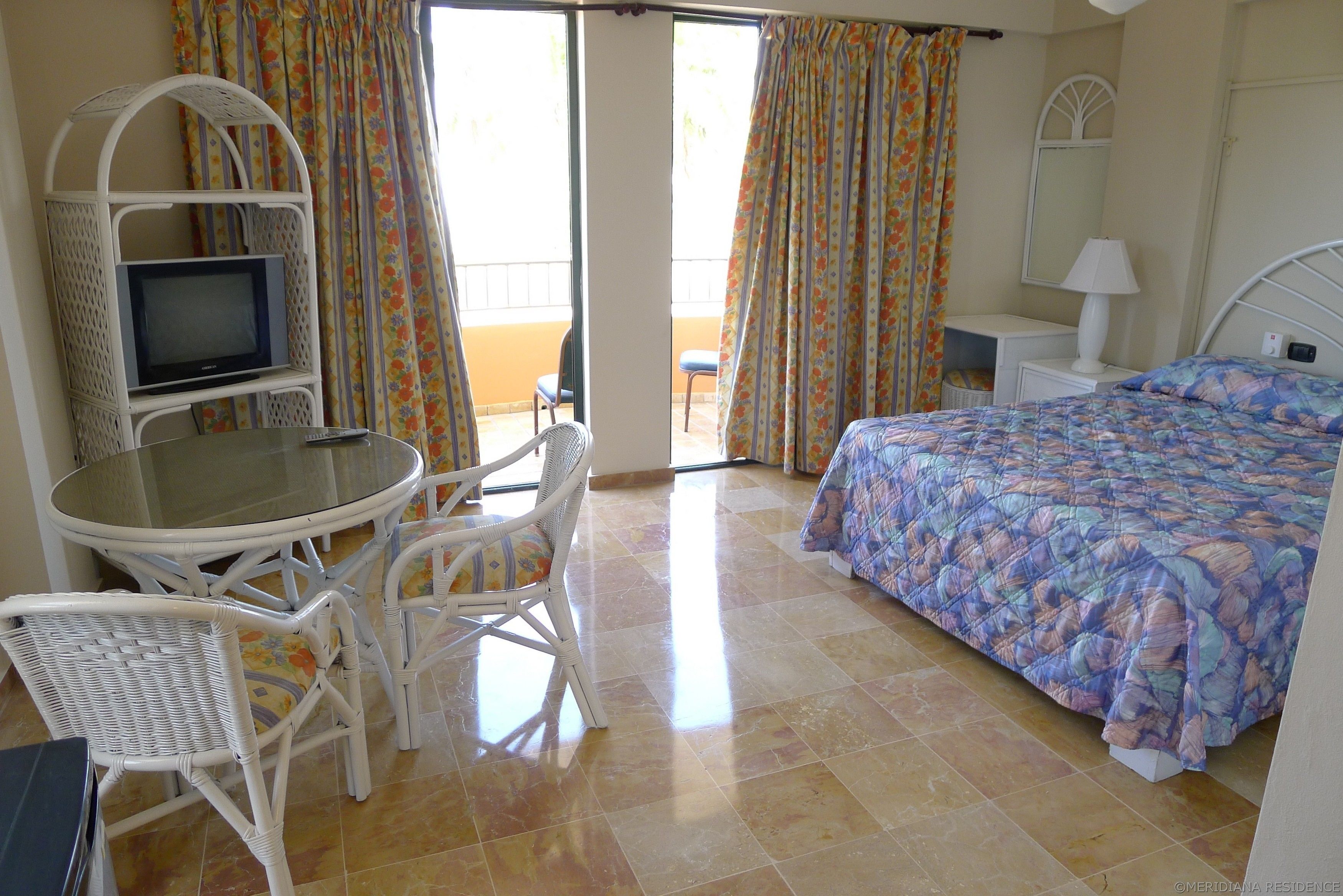 Une chambre d’hôtel comprenant un lit, un téléviseur posé sur un meuble, une table à manger avec des chaises et un balcon avec des rideaux.