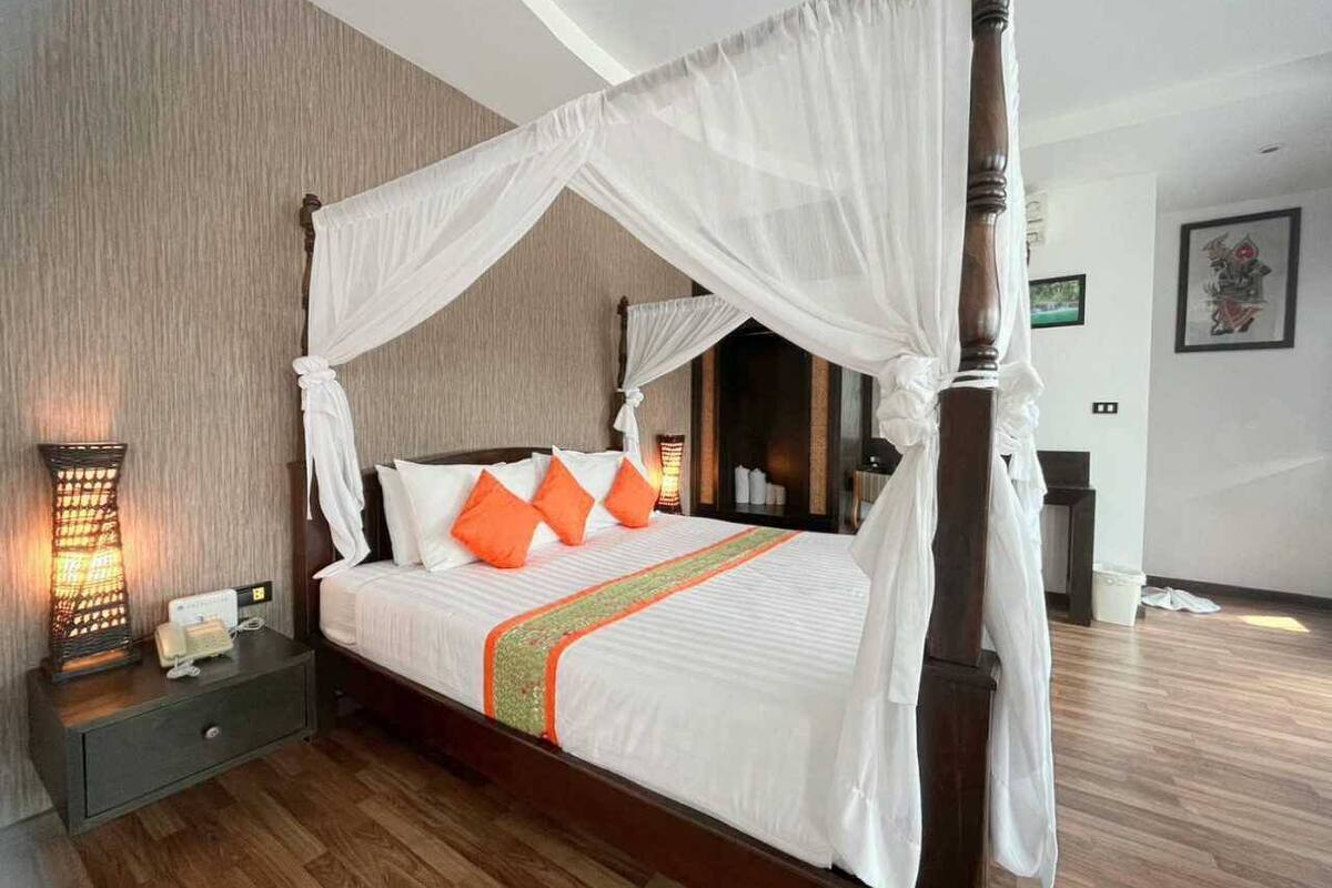 A four-poster bed with white linens and orange pillows, a bedside table with a lamp, and a wooden floor.
