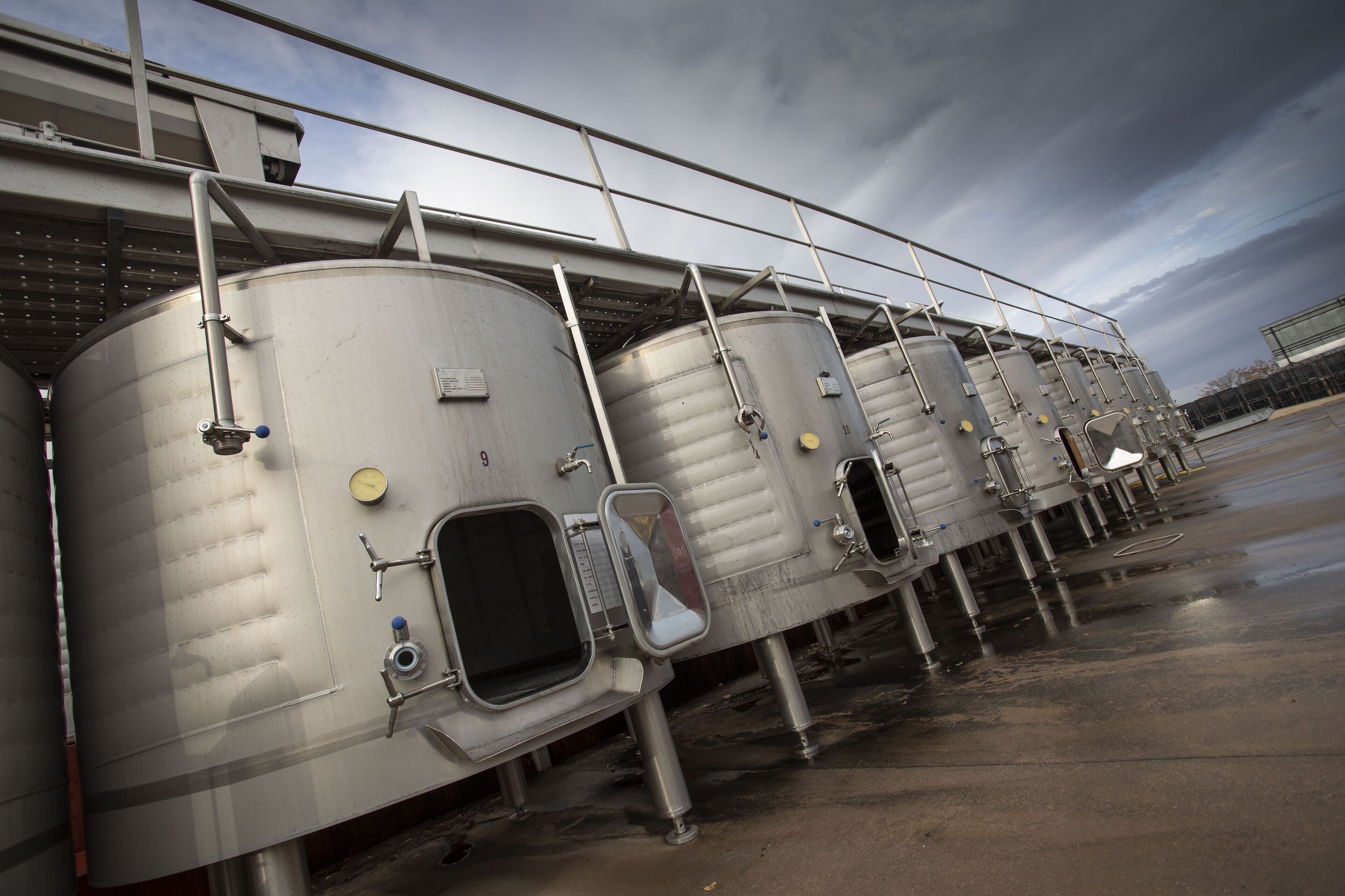 Una hilera de tanques de fermentación de acero inoxidable con indicadores de medición y válvulas, conectados por una pasarela metálica.