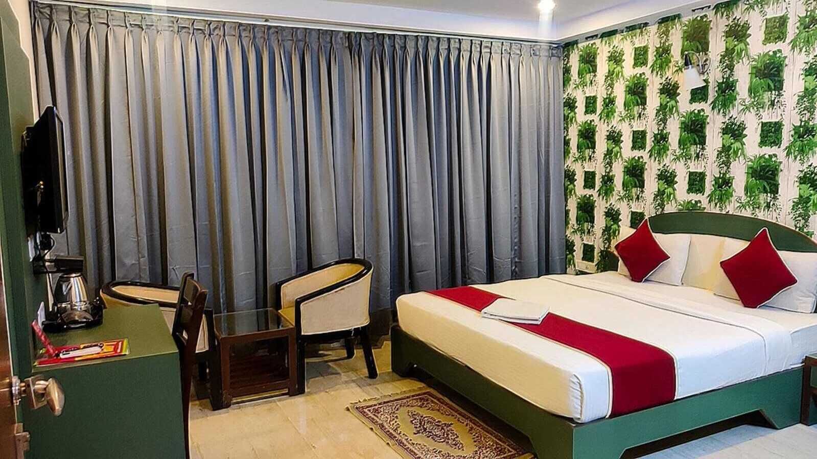 A hotel room with a bed, a desk, a chair, a television, and a wall with a green leafy pattern.