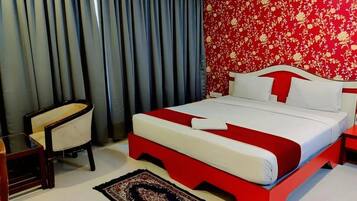 A hotel room with a bed, a chair, a nightstand, and a red floral wallpaper.