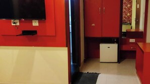 A compact room with a red wall, a small white refrigerator, a wooden cabinet, and a flat-screen TV mounted on the wall. - Royal Shades (Bengaluru)