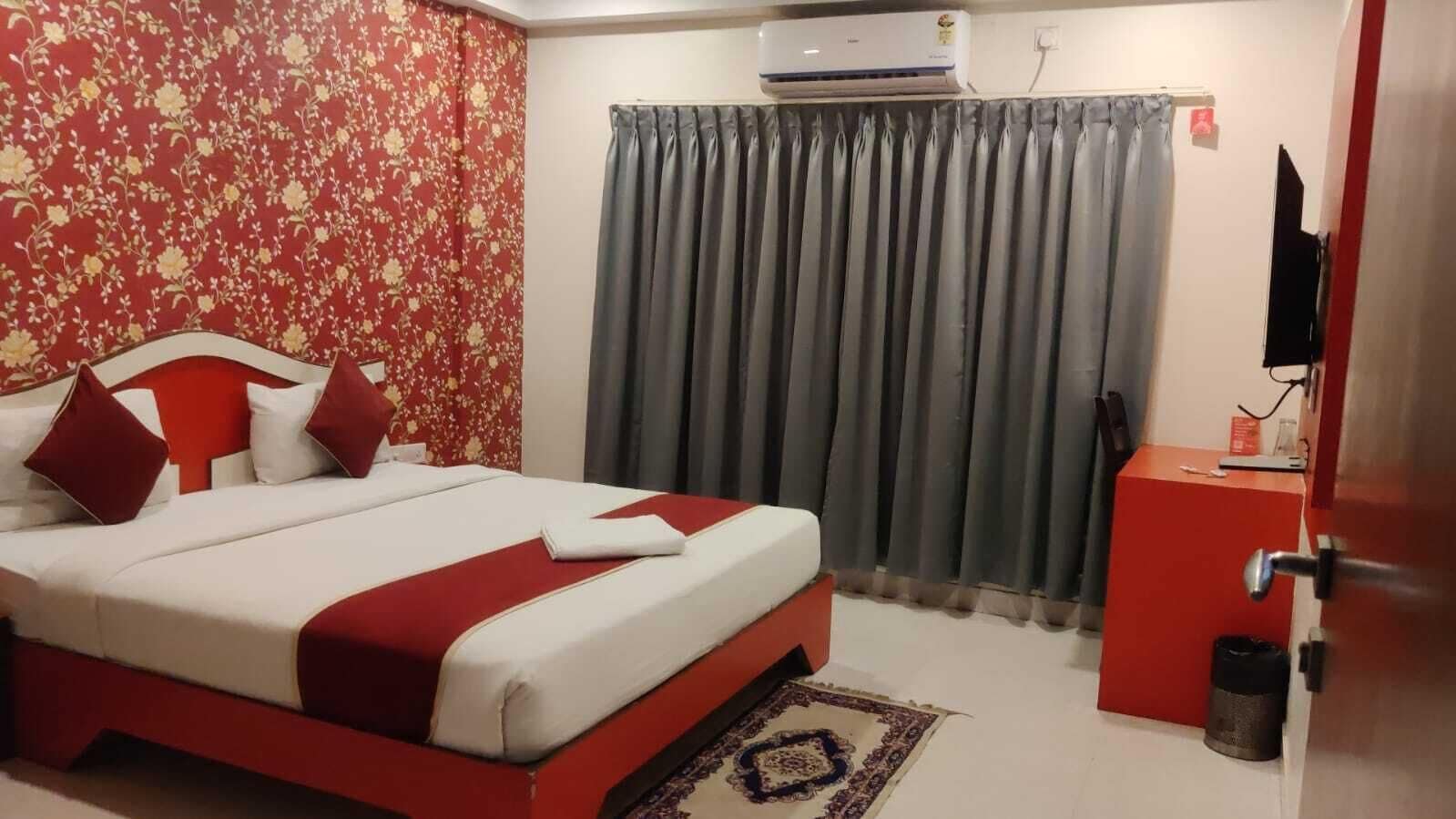 A hotel room with a bed, a red and white color scheme, a grey curtain, an air conditioning unit, a television, and a small desk with a trash bin.
