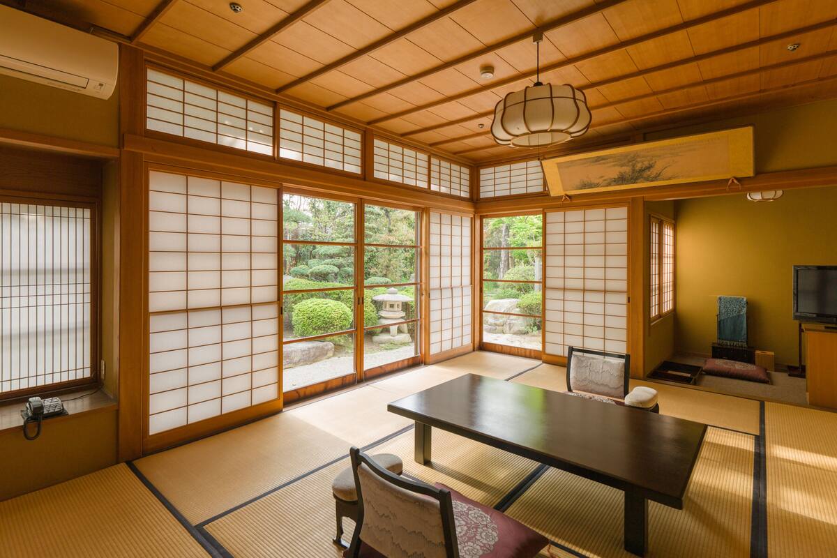 A traditional Japanese room with tatami flooring, a low table, a cushioned chair, sliding shoji doors, and a view of a garden.