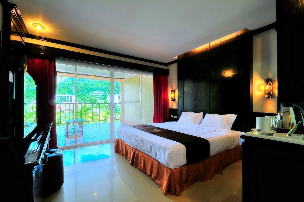 A modern hotel room with a large bed, a desk with a lamp, and a view of a green outdoor area through a sliding glass door.