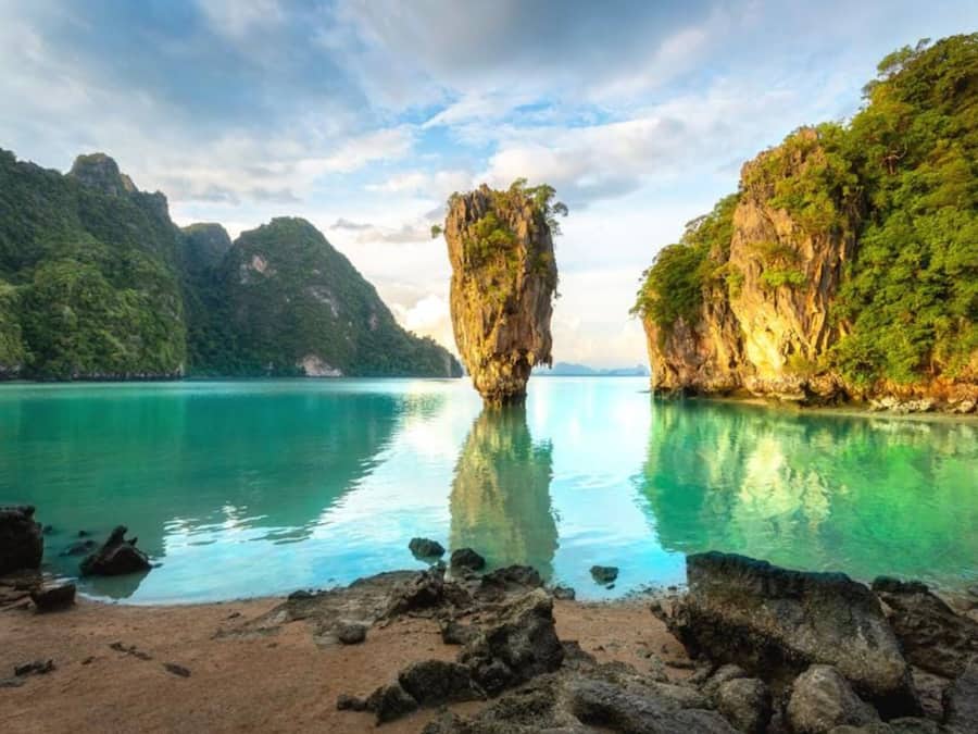 Un paisaje costero con una formación rocosa destacada en el agua, rodeado de vegetación exuberante y un cielo despejado.