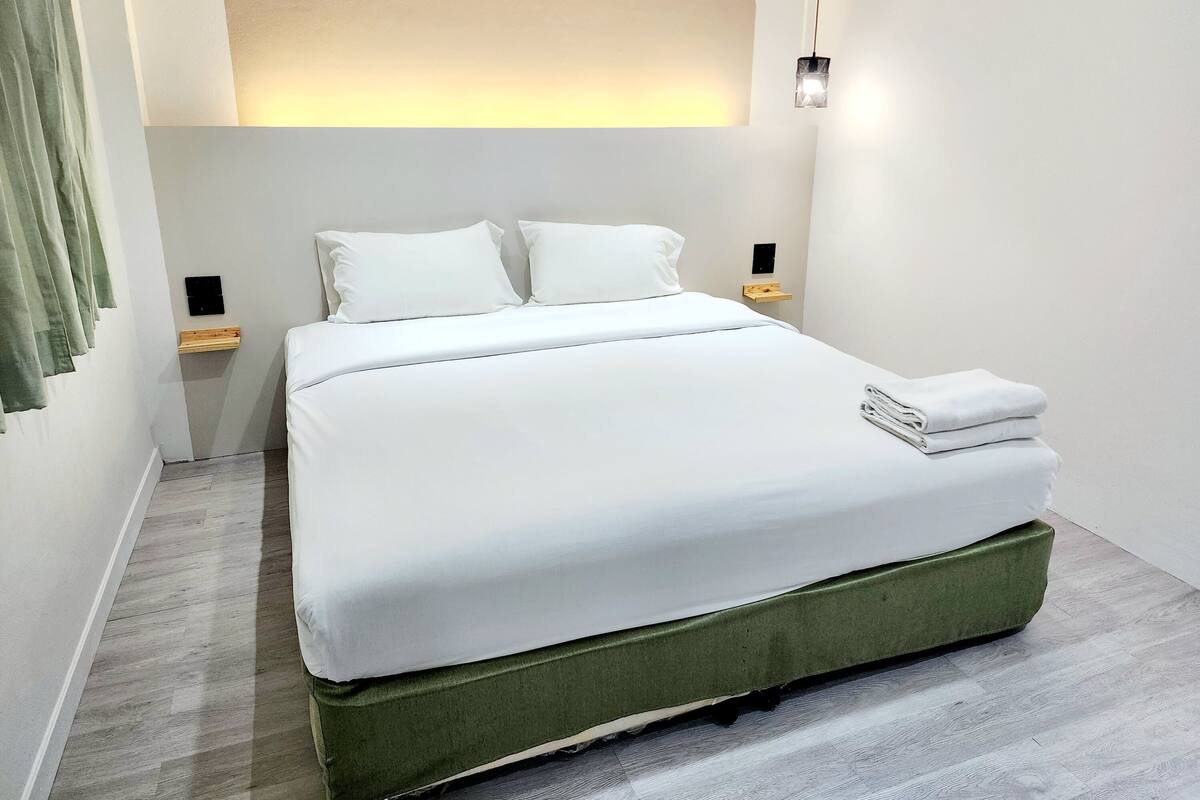 A neatly made bed with white linens in a modern room with a wooden floor and a green upholstered headboard.
