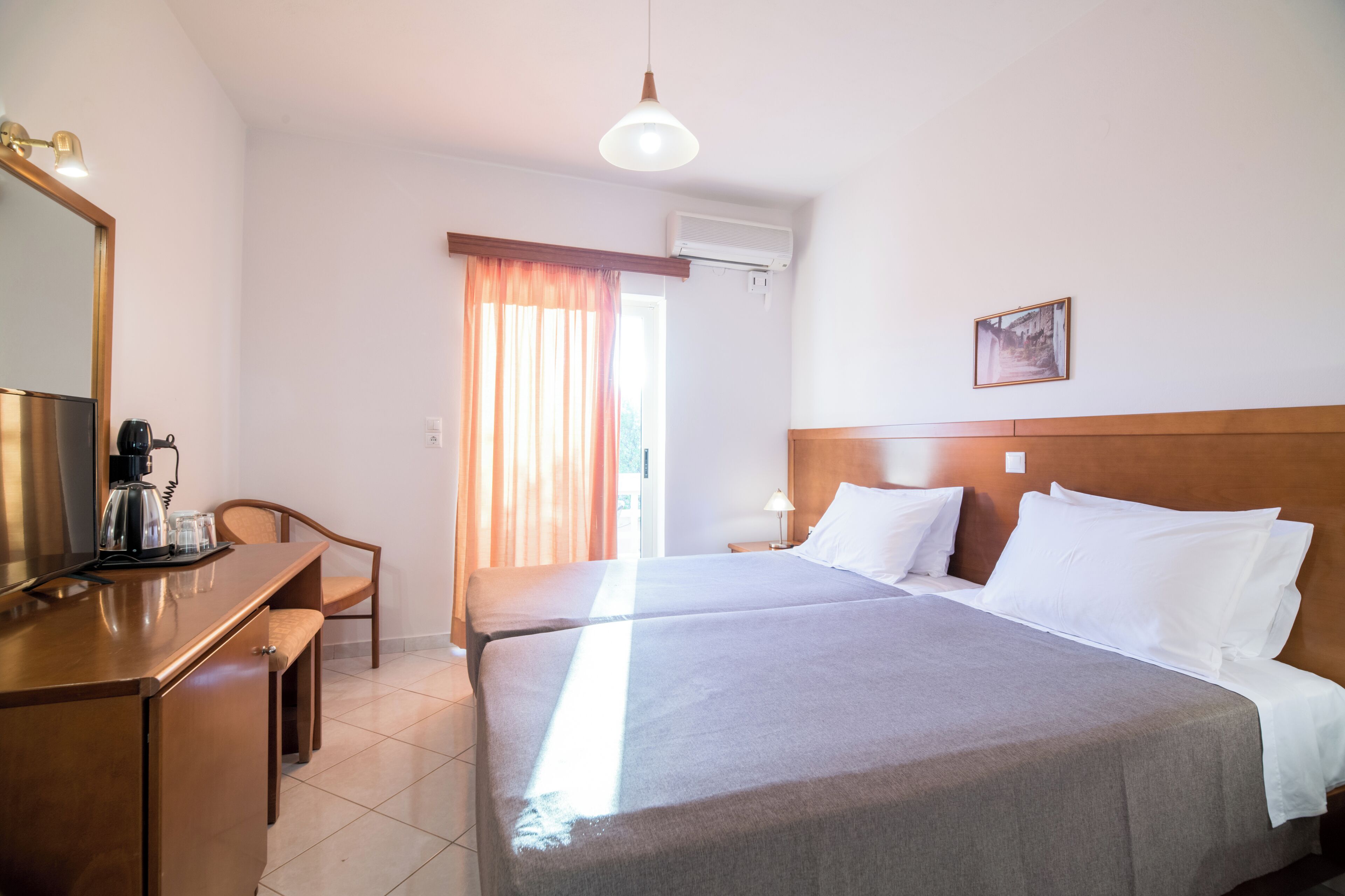 A hotel room with a bed, a desk with a coffee maker, a chair, a window with curtains, and a wall-mounted air conditioner.