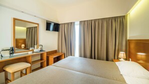 Quarto de hotel com cama, escrivaninha com cadeira, televisão e janela com cortinas.