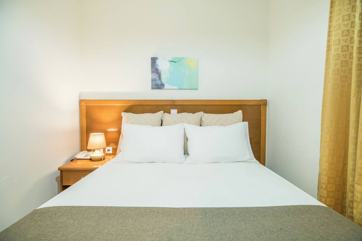A hotel room with a bed, a nightstand with a lamp, a telephone, and a painting on the wall.