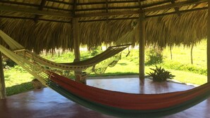 Una hamaca bajo una estructura de techo de paja, con piso de concreto y vista a un paisaje verde.