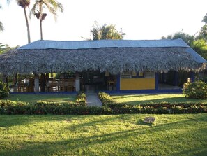 A thatched-roof building with a blue and yellow facade, surrounded by a well-maintained garden and palm trees. - Los Bohios Campo Añil (Jarabacoa)