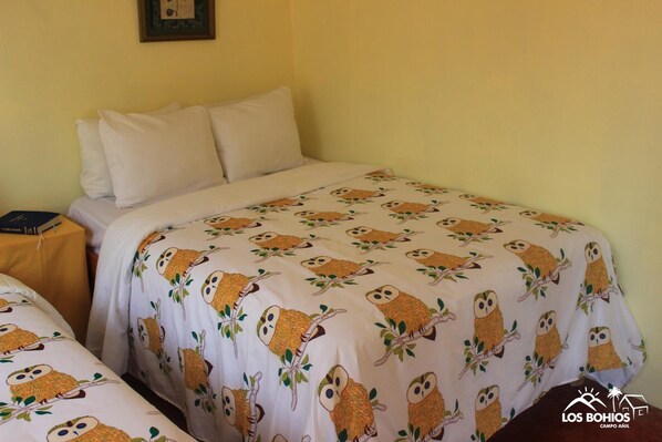 Una habitación con dos camas, cada una con una colcha blanca estampada con diseños de búhos. Hay una mesita pequeña con mantel amarillo y un cuadro en la pared.