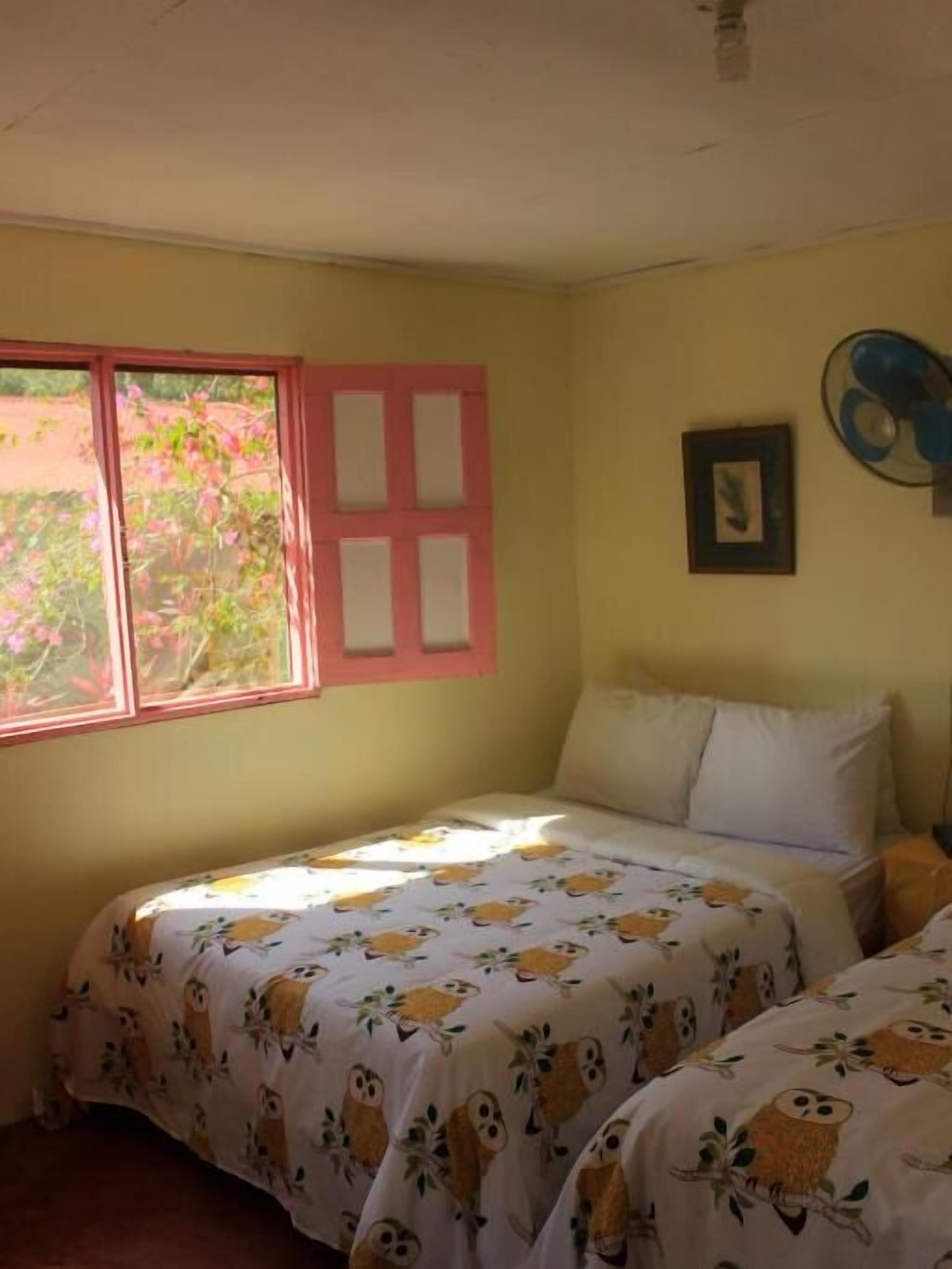 Una habitación con dos camas, una ventana con marco rosa, un ventilador de pared y un cuadro enmarcado.