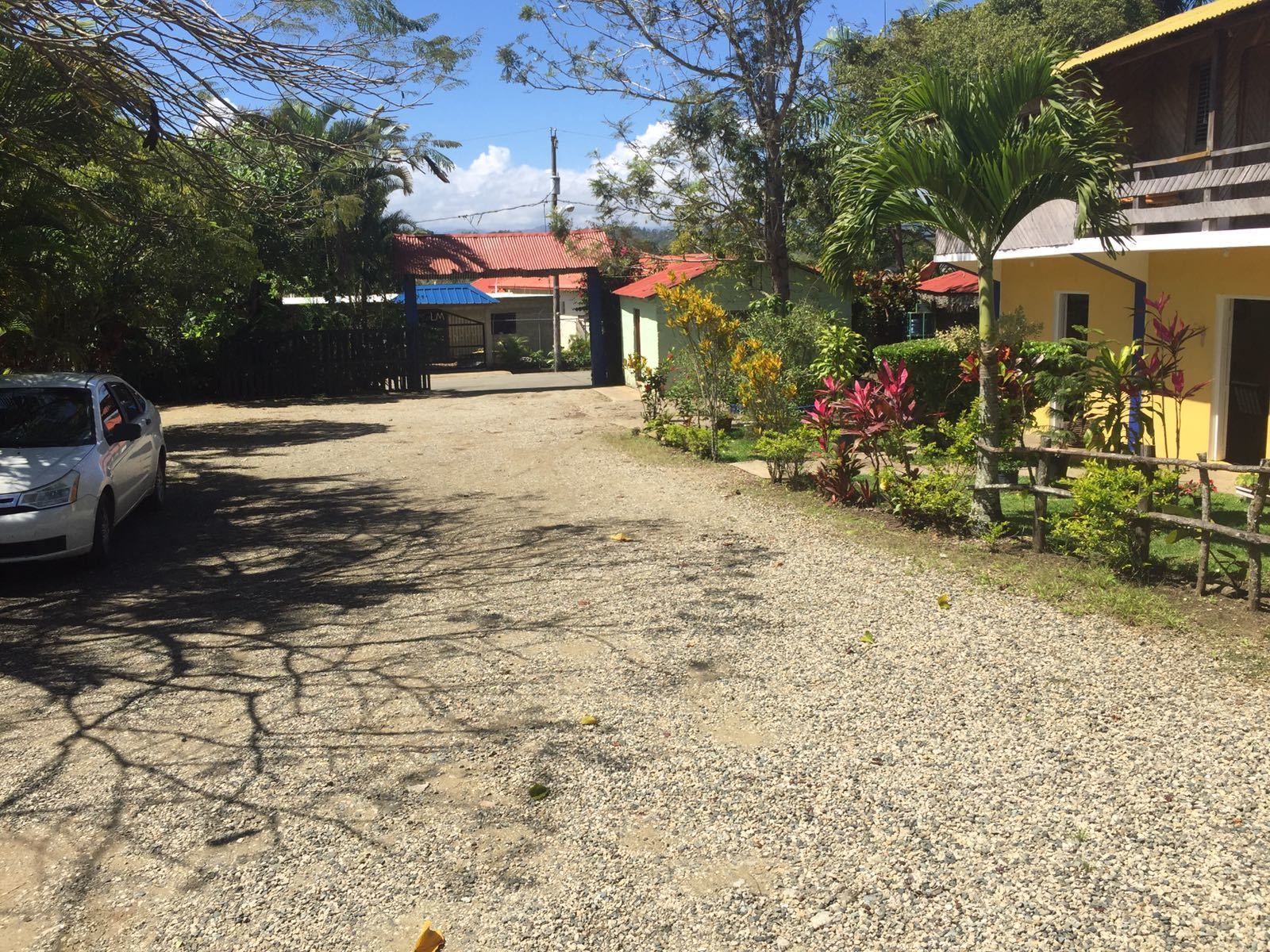 A gravel driveway leading to a building with a red roof, surrounded by greenery and a parked car.