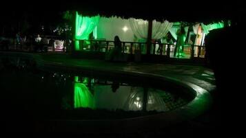 Espacio exterior iluminado con luces verdes, piscina y zona cubierta para sentarse.