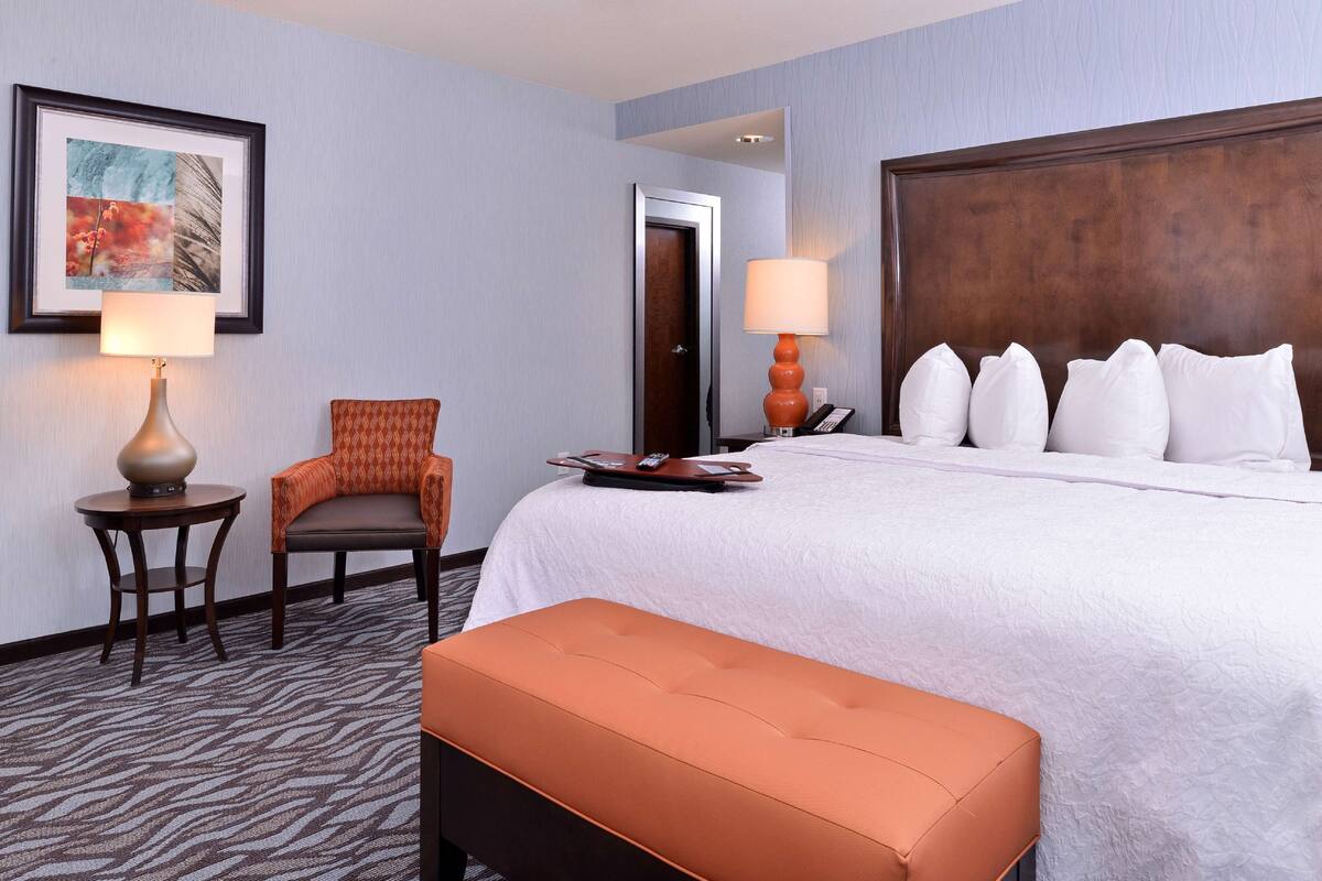 A hotel room with a large bed, a chair, a small table, and a wall-mounted lamp.