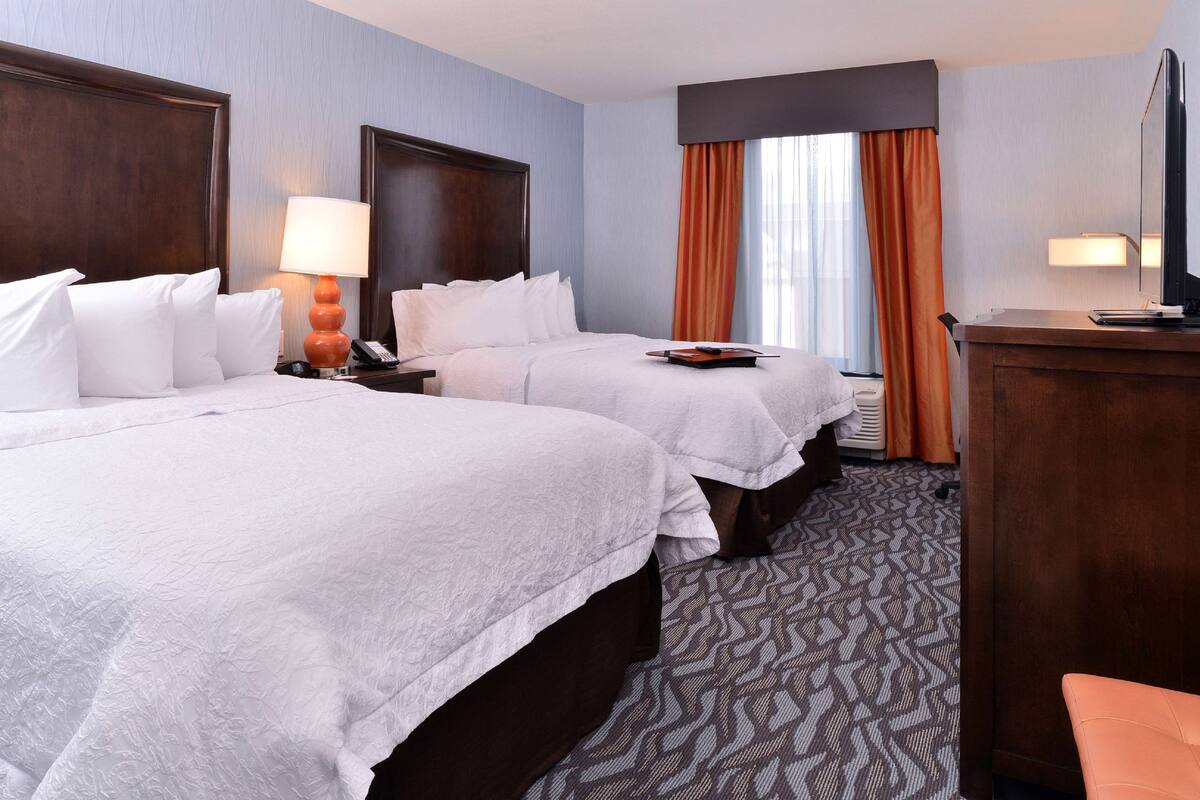A hotel room with two beds, a desk, a chair, a TV, and a window with curtains.