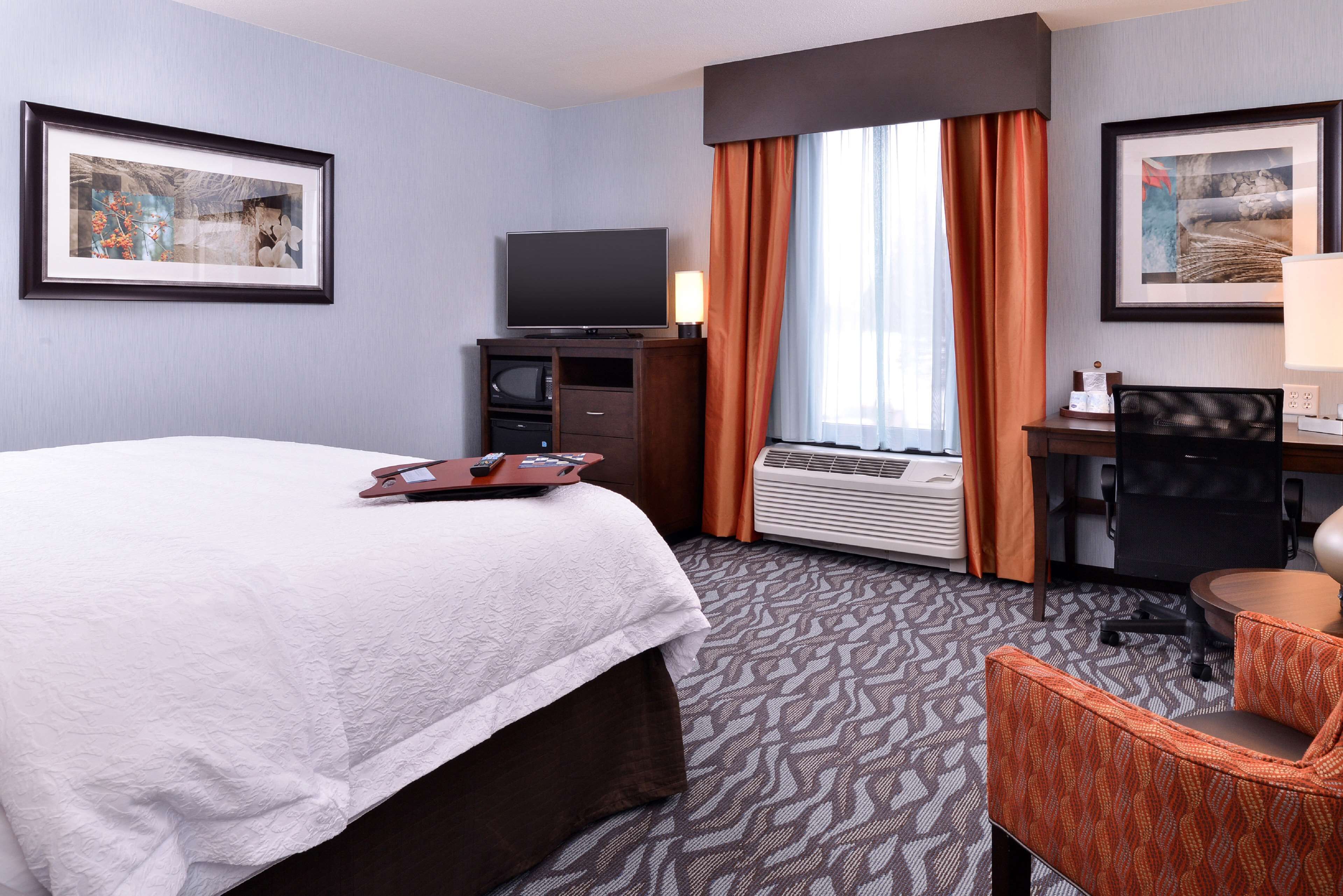 A hotel room with a bed, a desk with a chair, a television, and framed artwork on the walls.