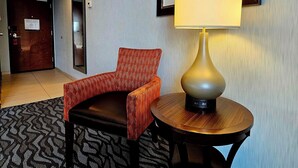 A hotel lobby with a patterned carpet, a red patterned chair, a round wooden table with a lamp, and a framed artwork on the wall.