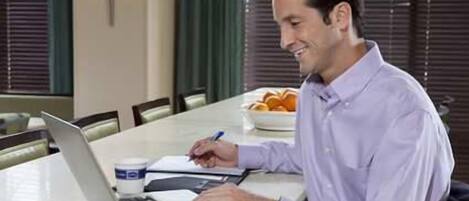 A person sitting at a table with a laptop, writing in a notebook. There is a bowl of fruit on the table.