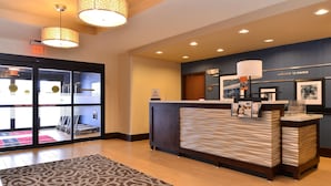 A hotel reception area with a modern design, featuring a reception desk, a 'welcome to Rome' sign, and a glass door entrance leading to a fitness area.