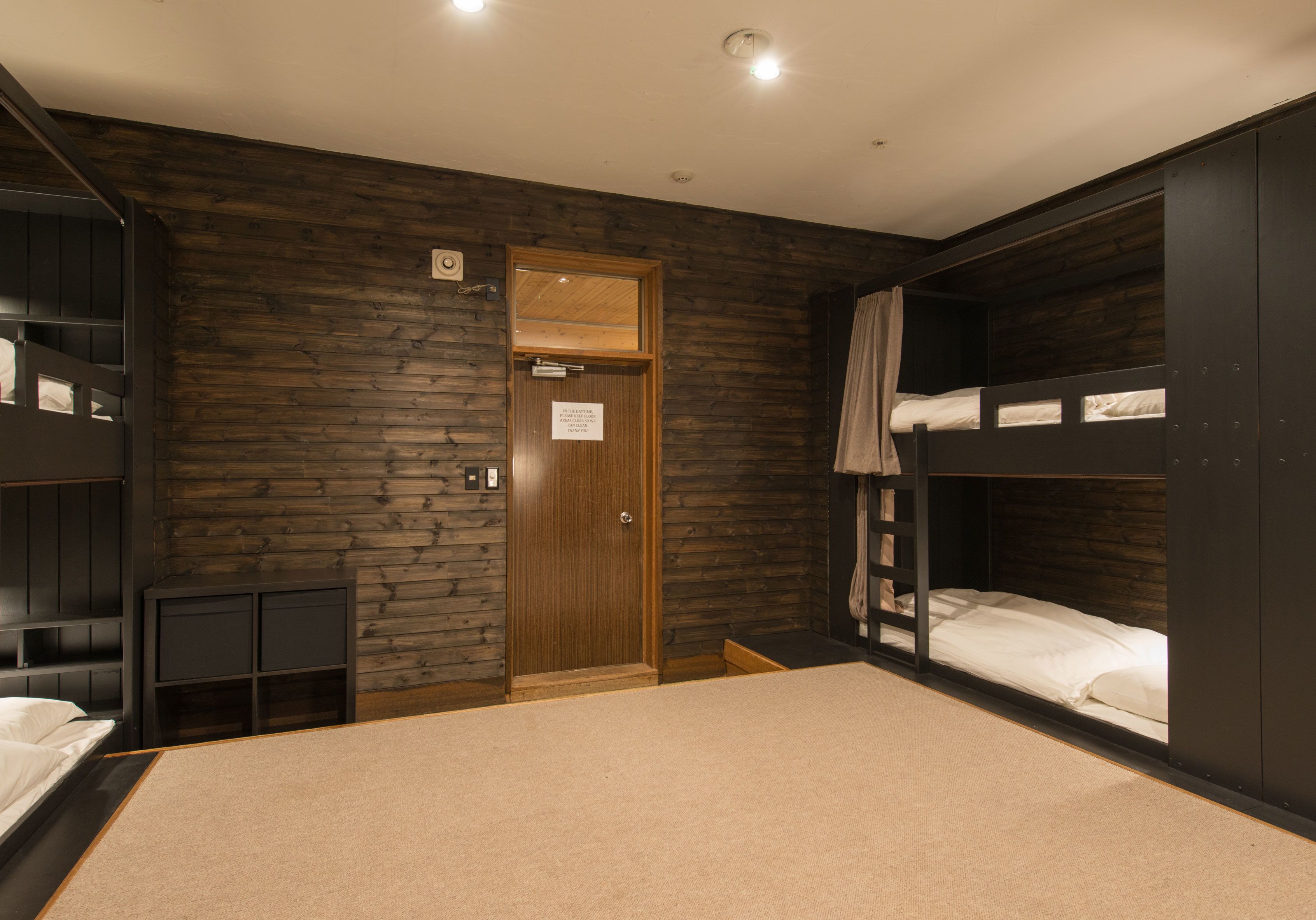 Een kamer met stapelbedden, een houten wand, een deur en een klein kastje.