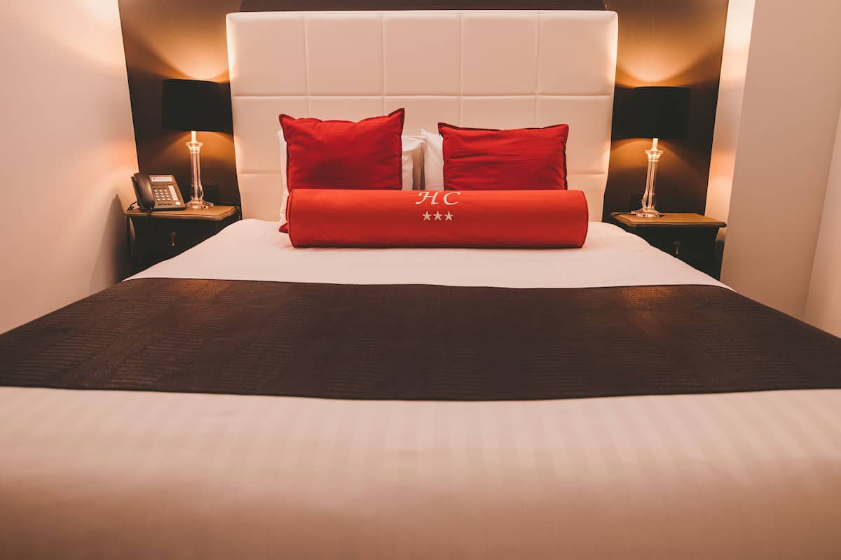 A neatly made bed with a white headboard, red pillows, and a red blanket with a logo. There are two bedside tables with lamps and a phone on one of them.