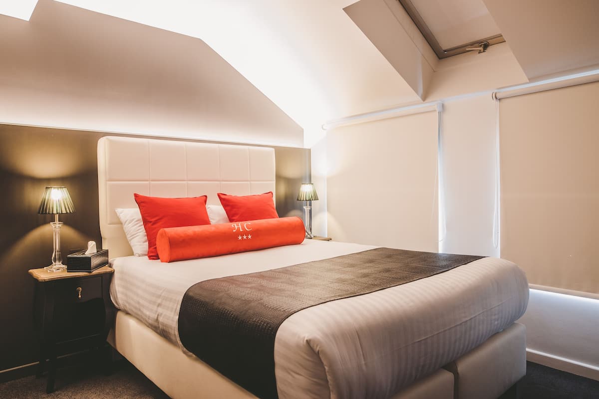 A hotel room with a large bed, a nightstand with a lamp, and a window with blinds.