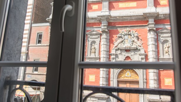 A view through a window with a metal railing, featuring a building with a red facade, white columns, and a central doorway with a decorative arch. - Hôtel Hors Château (Liege)