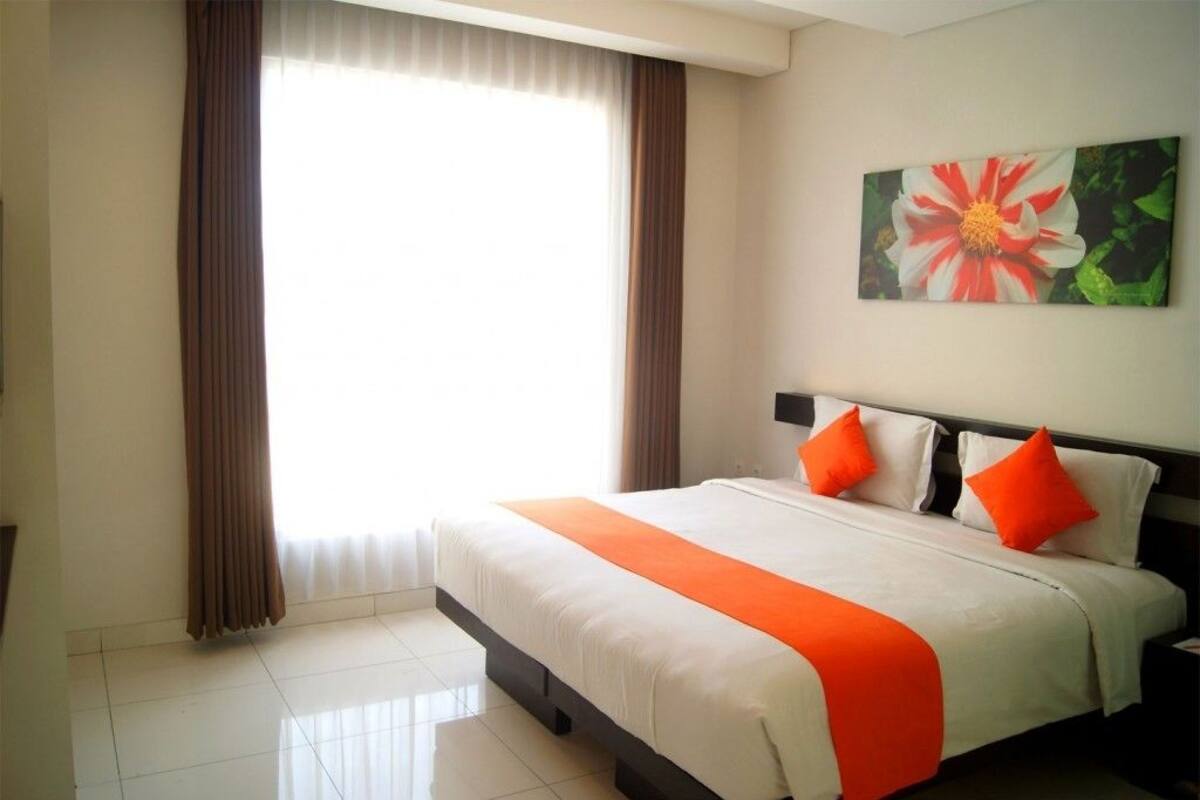 Ein Hotelzimmer mit einem großen Bett, braunen Vorhängen, einer weißen und orangefarbenen Bettdecke und einem Blumenbild an der Wand.