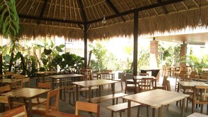 An outdoor dining area with wooden tables and chairs, surrounded by lush greenery and a thatched roof structure.