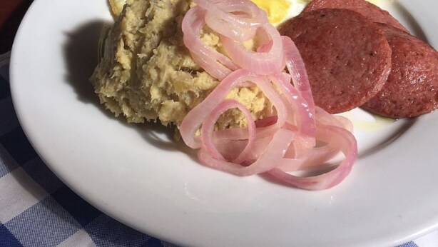 A plate with scrambled eggs, a slice of ham, pickled onions, and a piece of bread.