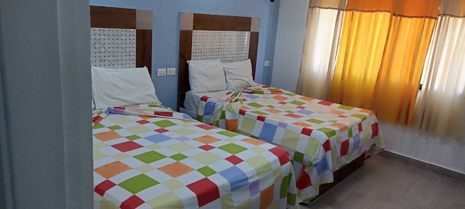 A room with two beds, each with a colorful checkered bedspread and white pillows. The room has a simple headboard design and a window with curtains.