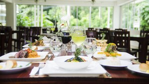 A dining table set with various dishes, glasses, and a bottle of wine in a restaurant with large windows and a view of greenery. - Hotel Arenas en Punta Leona (Tárcoles)