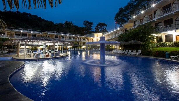 A swimming pool with a central fountain, surrounded by lounge chairs and a building with multiple balconies. - Hotel Arenas en Punta Leona (Tárcoles)
