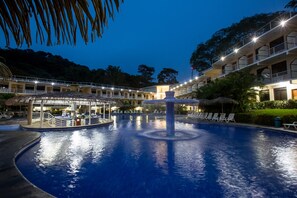 Piscina com uma fonte central, rodeada por espreguiçadeiras e um edifício com várias varandas.