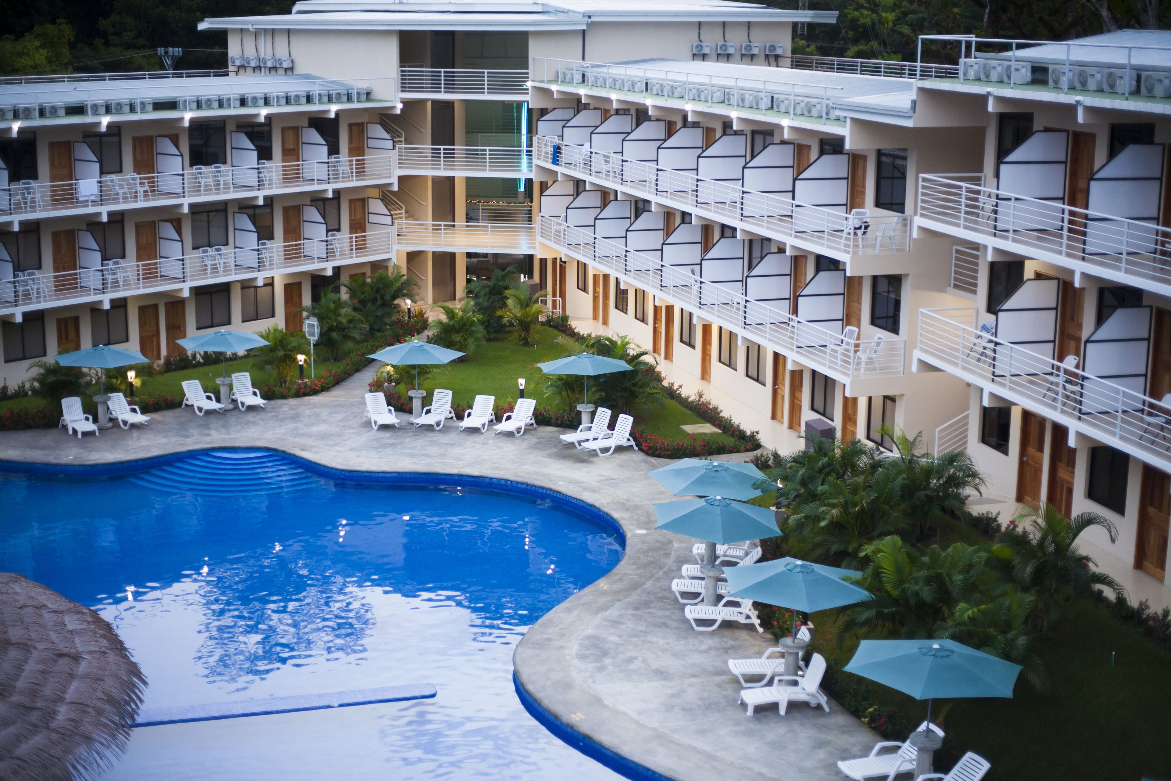 Un hotel de varios pisos con piscina central, sillas de descanso al aire libre y sombrillas.