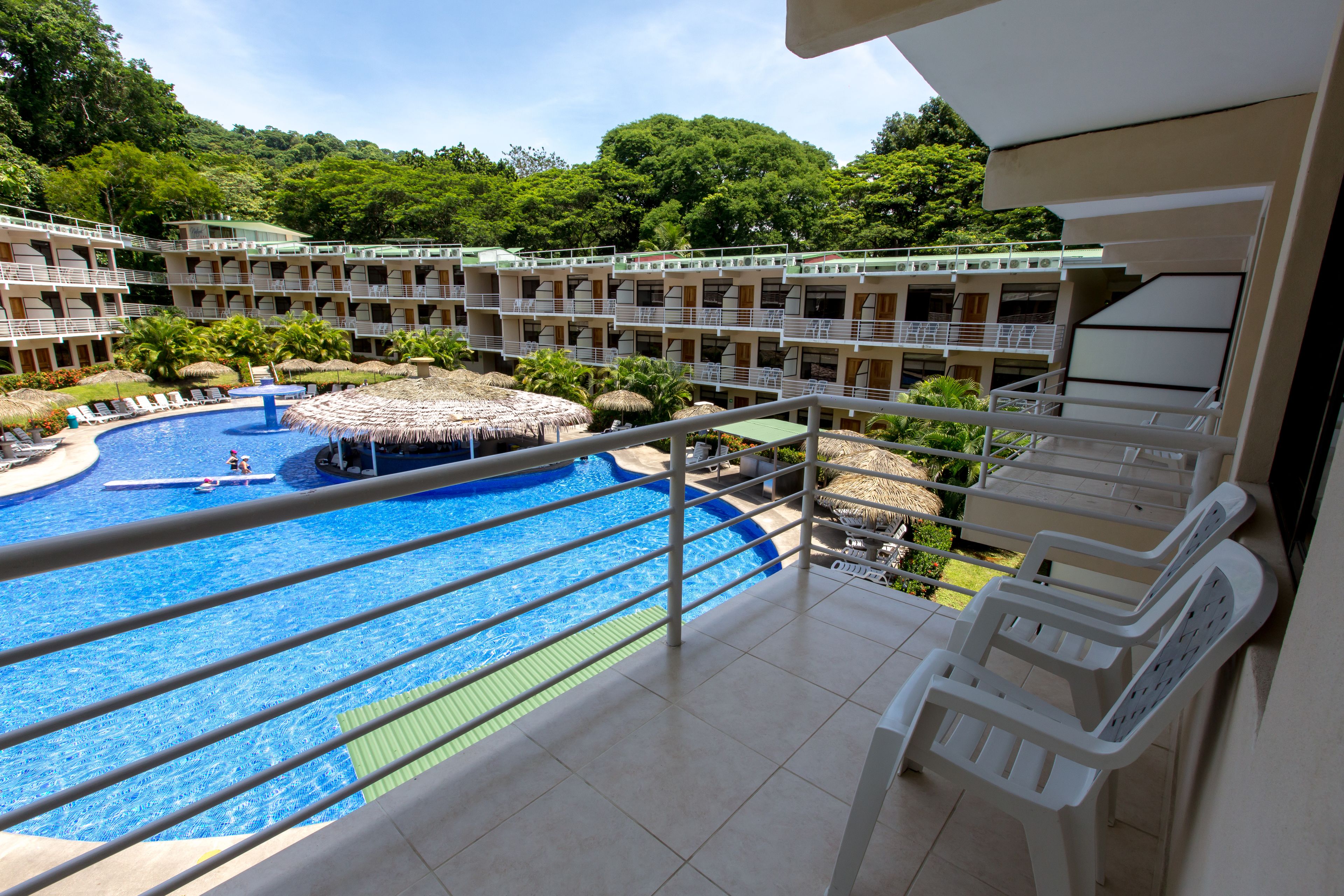 Un hotel con piscina, varios balcones y abundante vegetación al fondo.