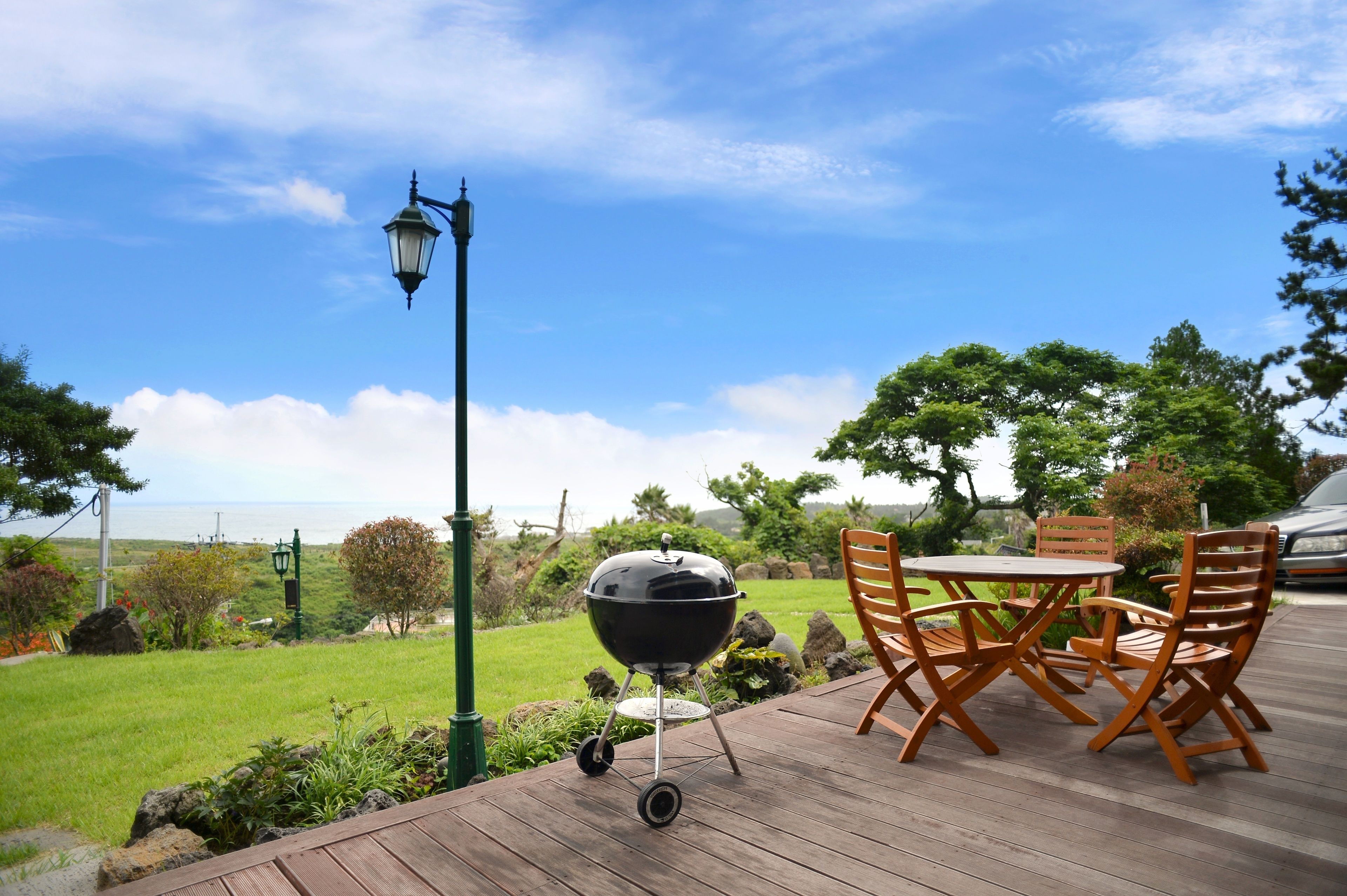 Una terraza de madera con una parrilla, una mesa y sillas exteriores, con vistas a un área verde con árboles y el océano.