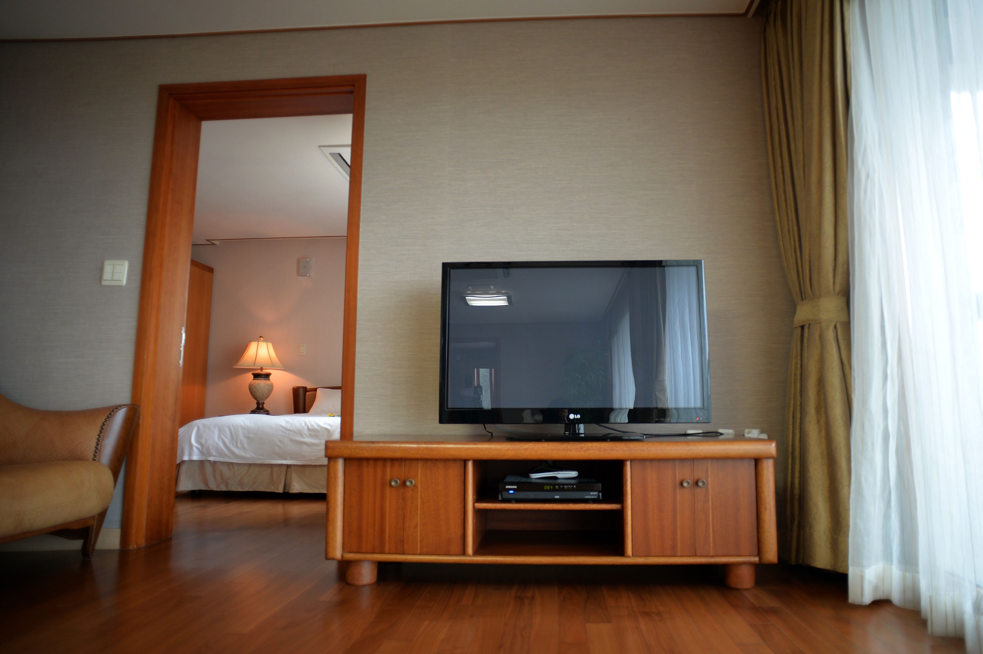 A hotel room with a wooden TV stand, a bed, a lamp, and a view of another room through an open door.