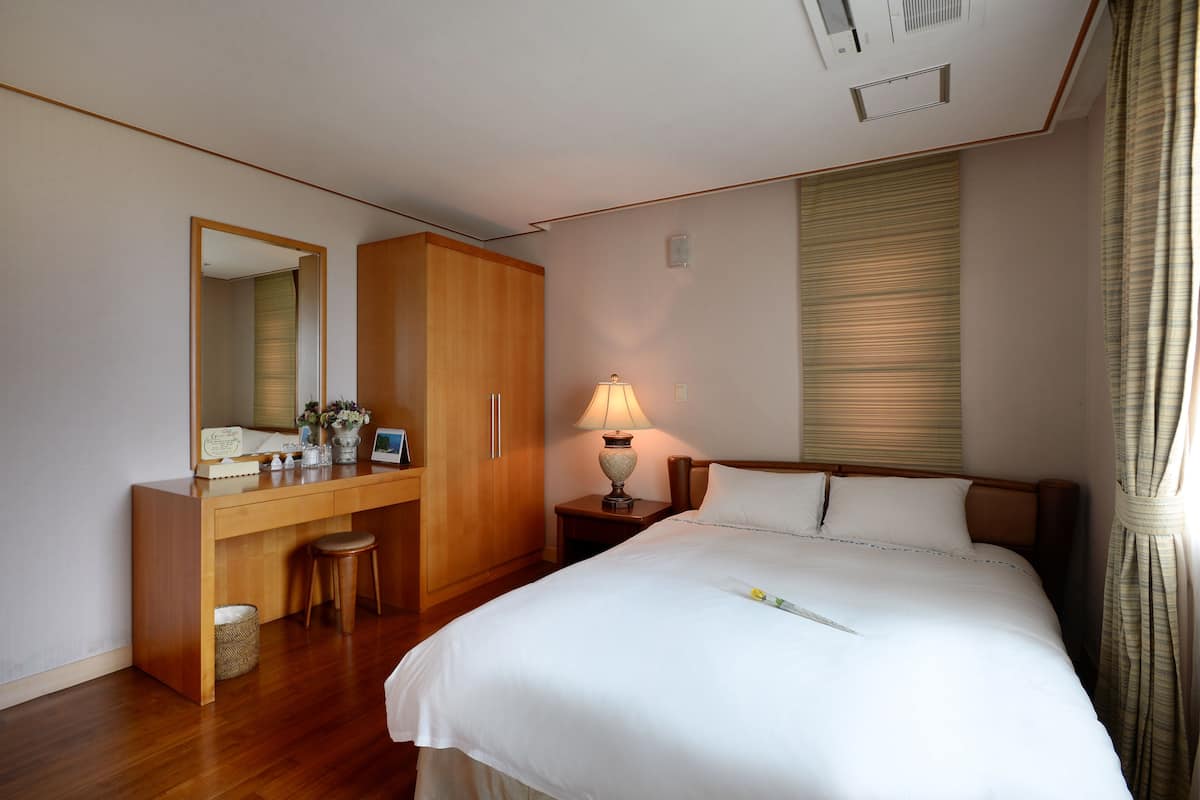 Une chambre d’hôtel avec un bureau en bois, un grand miroir, une table de chevet avec une lampe, un lit avec du linge de lit blanc et une fenêtre avec des stores.