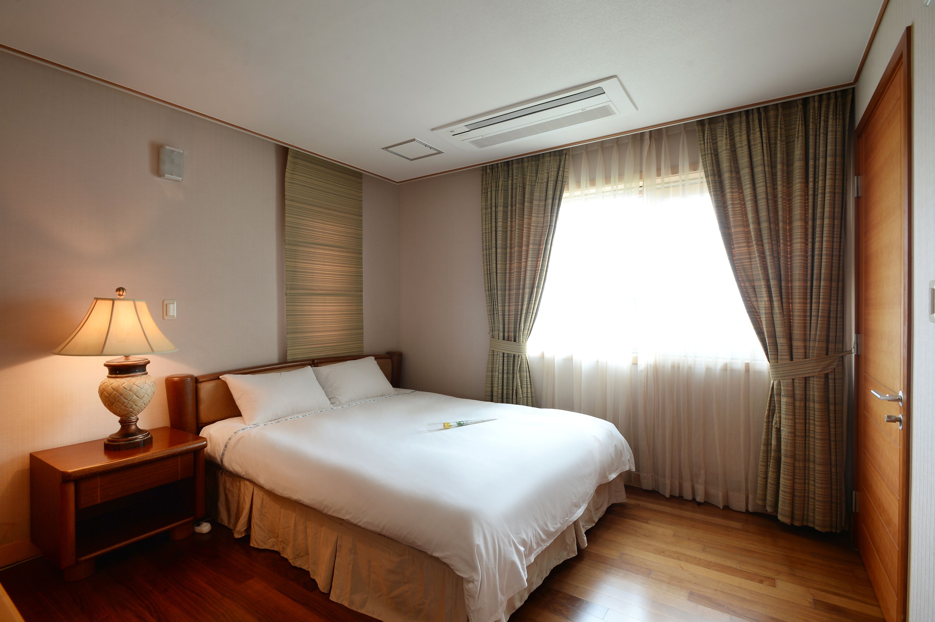 Una cama bien tendida en una habitación de hotel, con una mesita de noche, una lámpara y una ventana con cortinas.