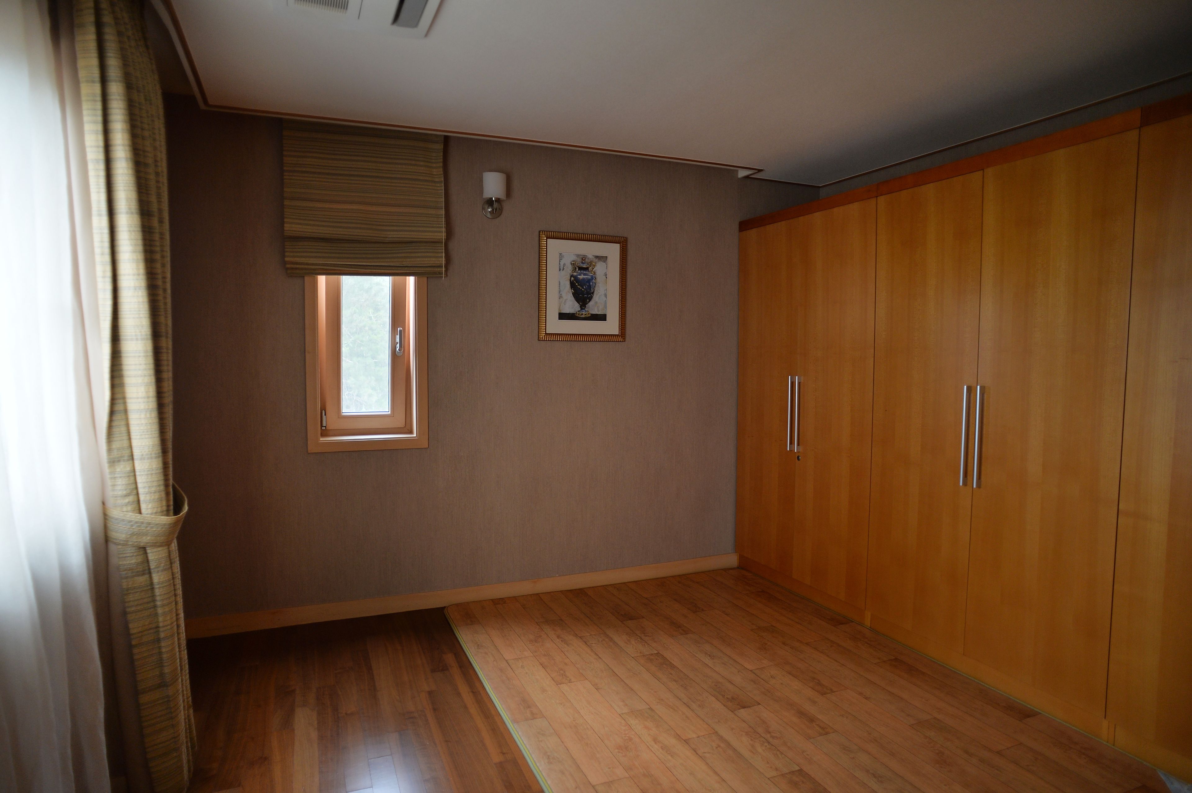 A room with wooden flooring, a window with blinds, a framed picture on the wall, and a wooden wardrobe.