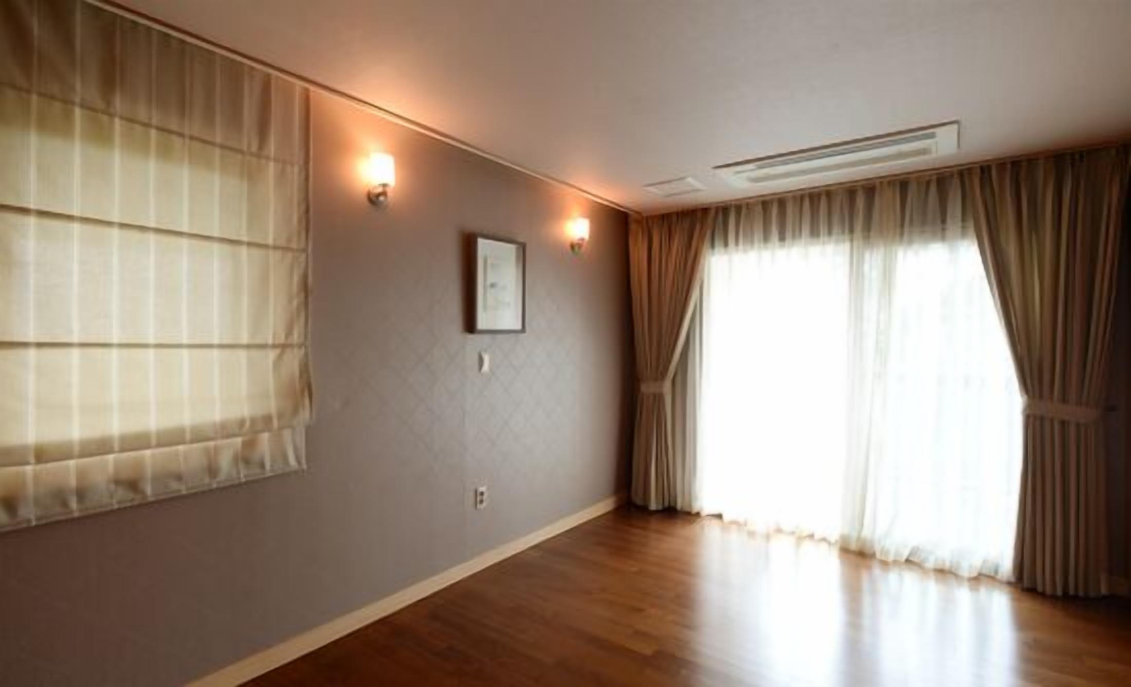 Ein Zimmer mit Holzboden, einem großen Fenster mit Vorhängen und zwei Wandleuchten.