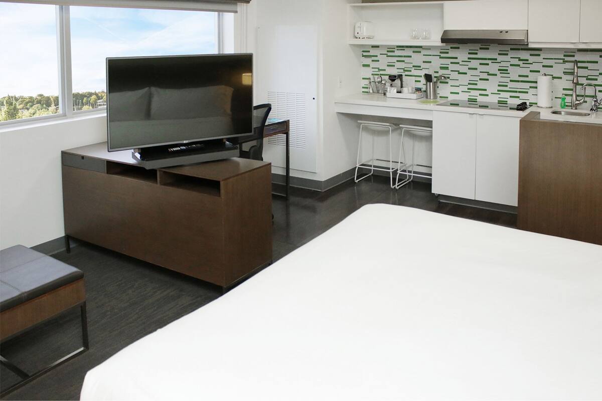 Habitación de hotel moderna con televisión de pantalla plana sobre un soporte de madera, cama blanca, pequeña mesa de madera, kitchenette con encimera blanca y salpicadero verde, y ventana con vista a la ciudad.