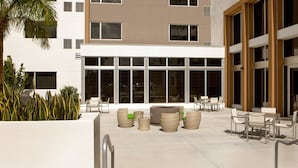 Un espacio exterior moderno con mobiliario de mimbre y una chimenea, rodeado de amplias ventanas y una palmera.