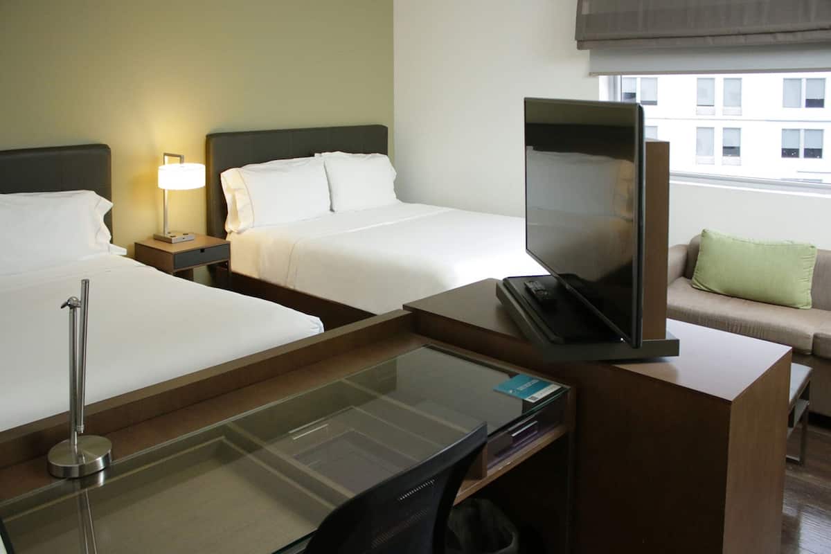 Habitación de hotel con dos camas, un escritorio con televisión de pantalla plana, una lámpara y un sofá con un cojín.