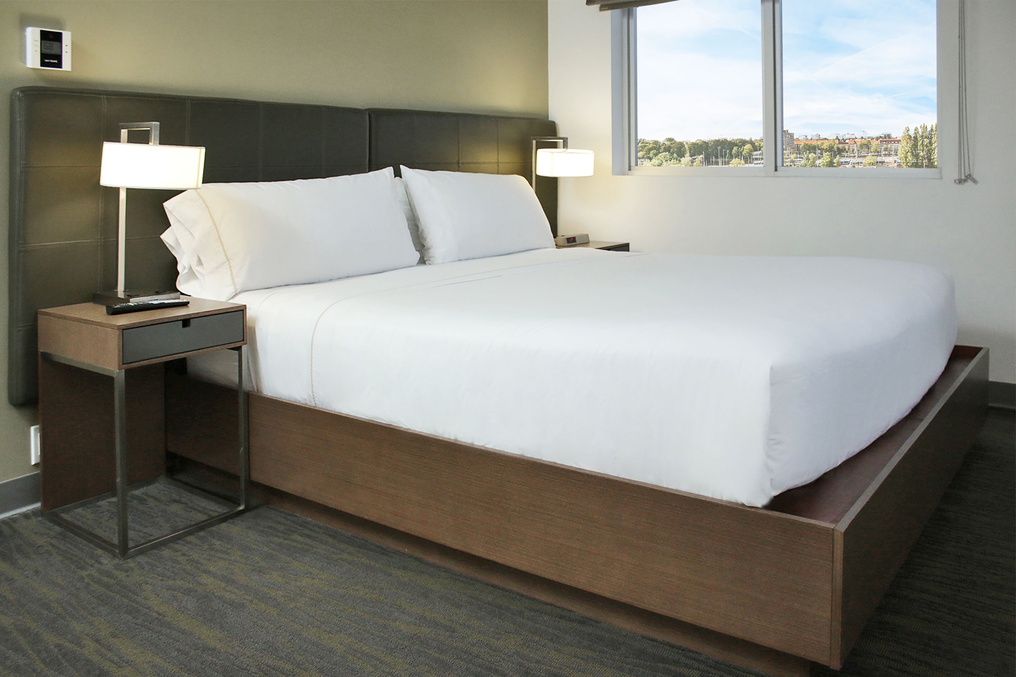 A hotel room with a large bed, two bedside tables with lamps, a view of the city through the window, and a control panel on the wall.